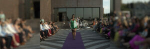 Image that shows a model wearing a bright green gown on a runway as part of Avivo's Mission & Mingle event. There's a play button on top, as this image is a button to play a recap of the event.